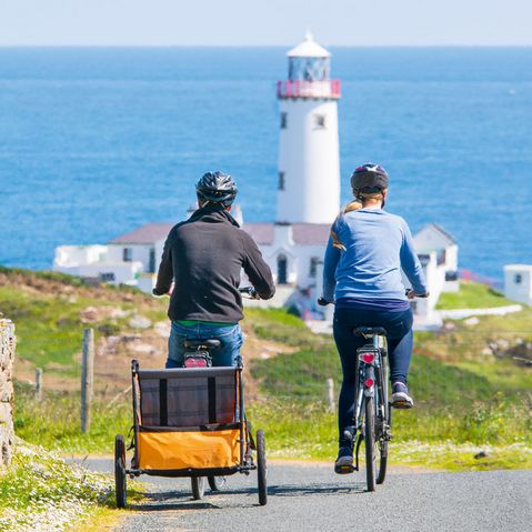 Zwei Radfahrer fahren an der Küste Irlands entlang, geradewegs auf einen weißen Leuchtturm zu